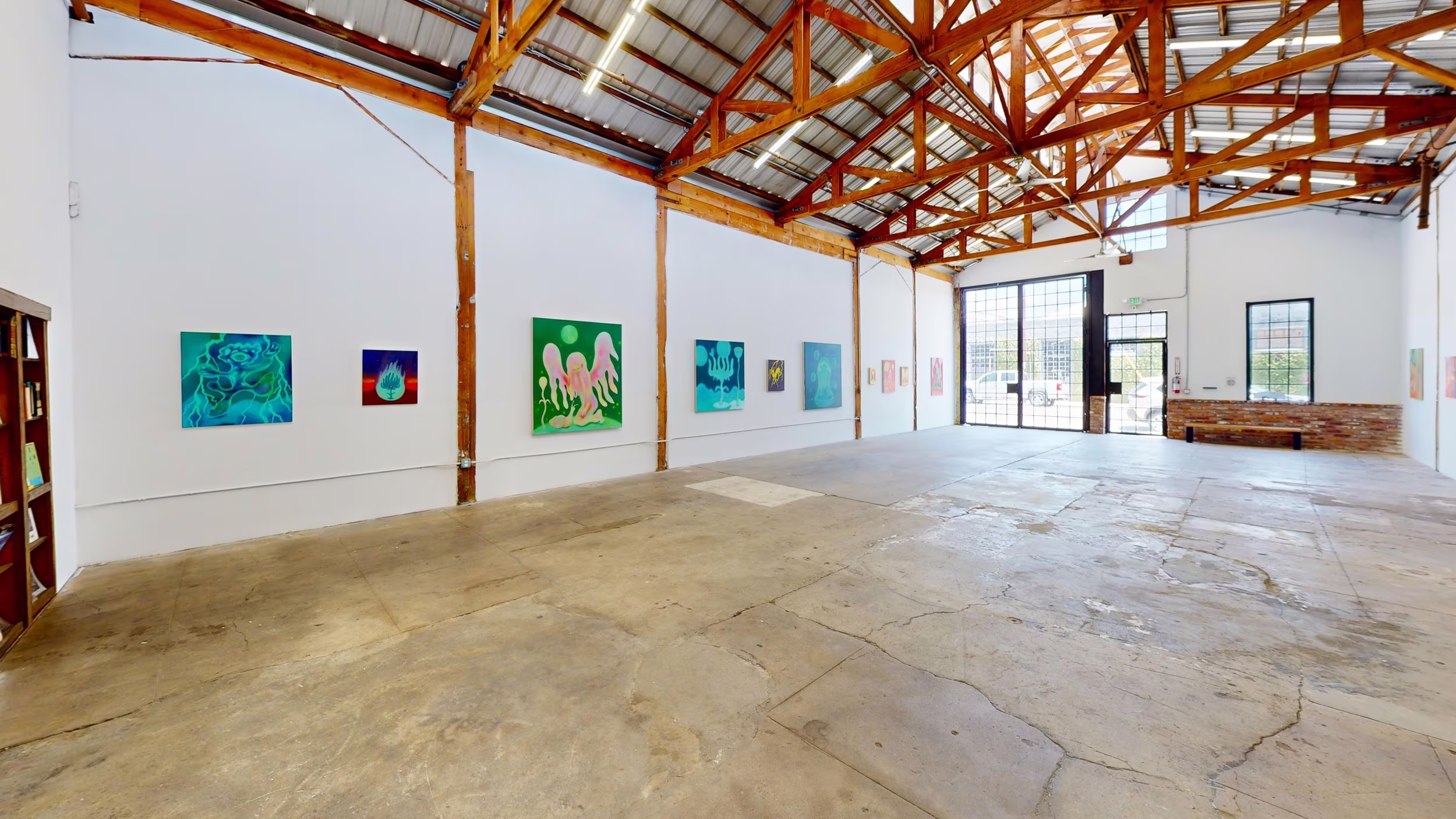 Open gallery with turquoise and pink paintings along both walls, concrete floor, and gridded glass entry.