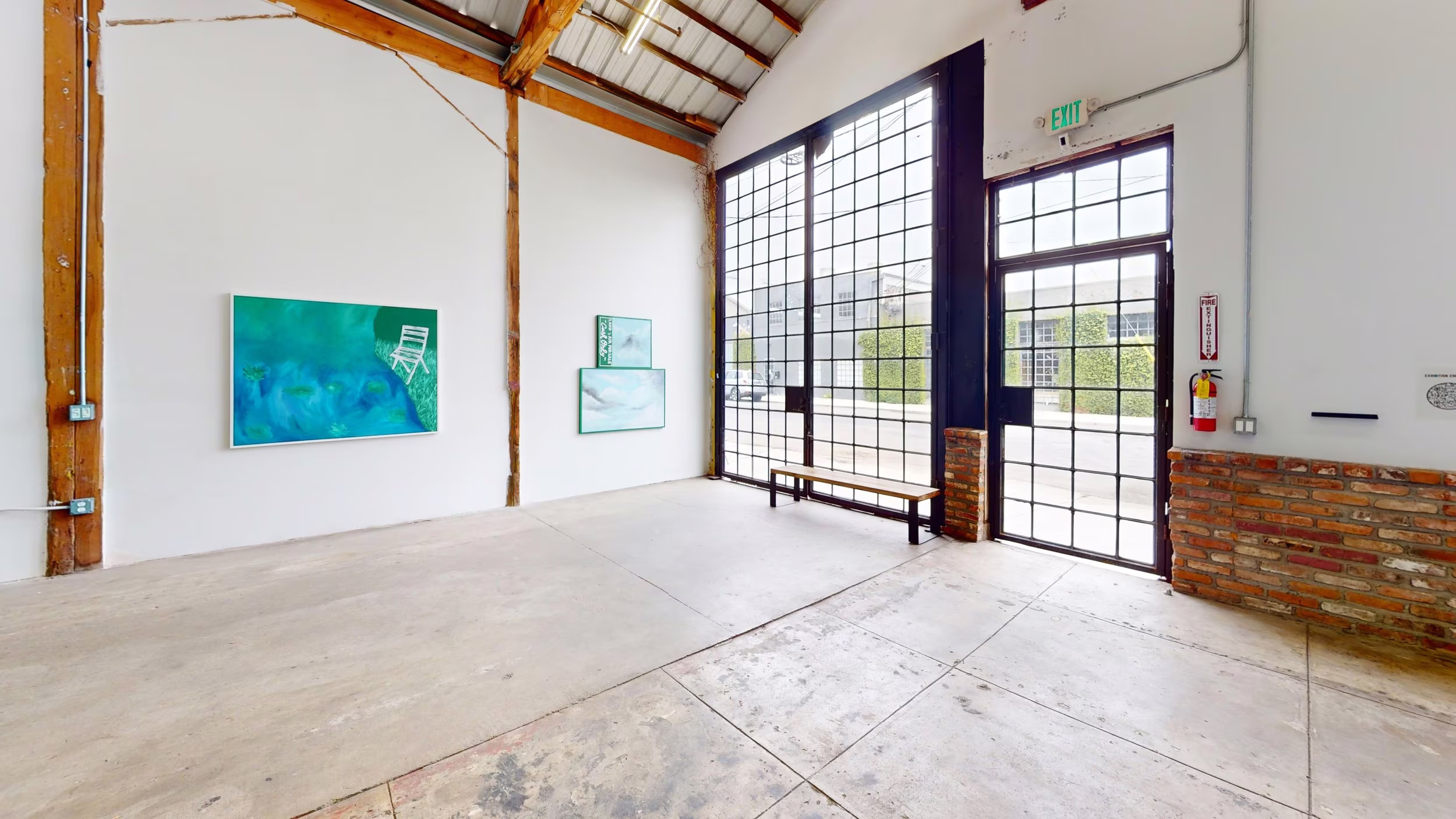 Bright storefront corner with gridded glass doors, exposed brick base, bench, and three cool-toned artworks.