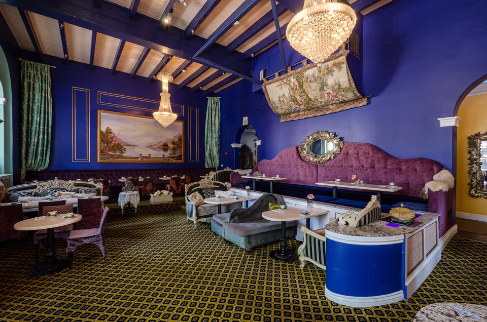 Reverse angle of the deep-blue dining room showing its long banquette, round café tables, chandeliers, and open floor area.