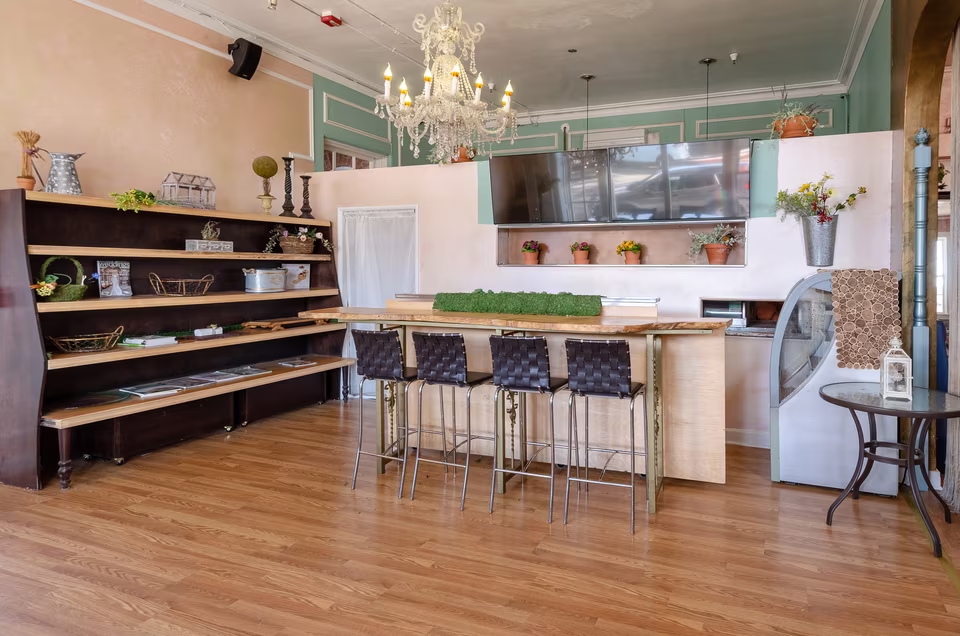 Pastel café counter with live-edge wood bar, stools, chandelier, display shelving, and wall-mounted screen.
