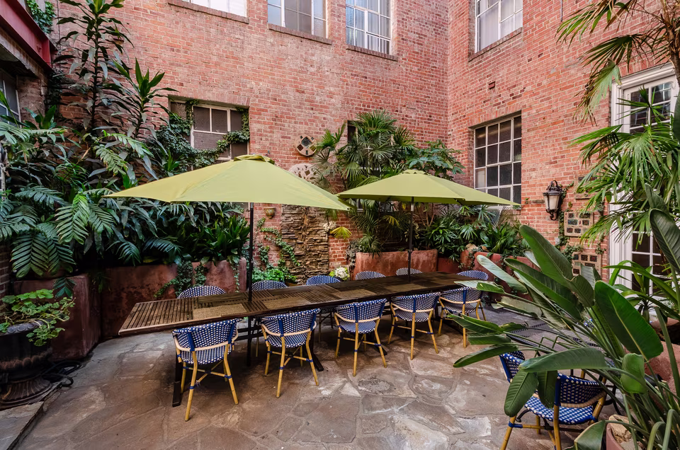Wide patio view with twin green umbrellas, brick walls, café seating, stone floor, and mature greenery.