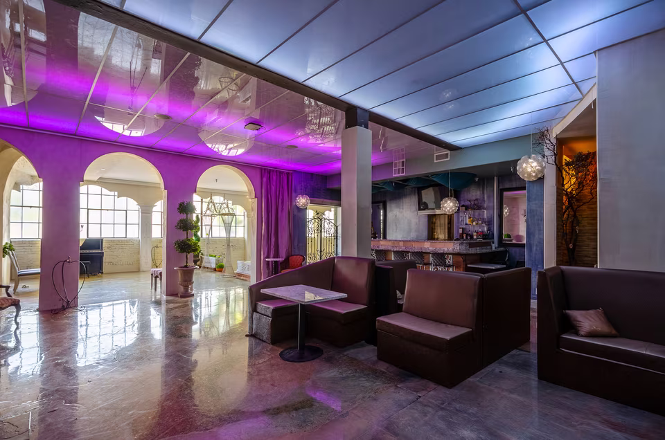 Reverse angle across the lounge toward tall arched windows, booth seating, bar, and reflective open floor.