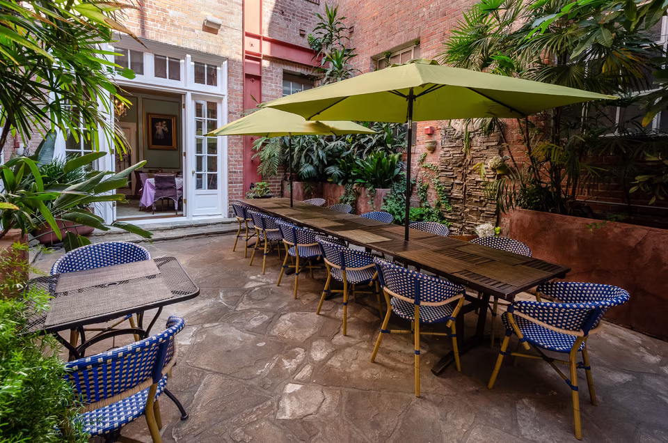 Leafy brick patio with long wood tables, blue woven chairs, green umbrella, and dense tropical planting.