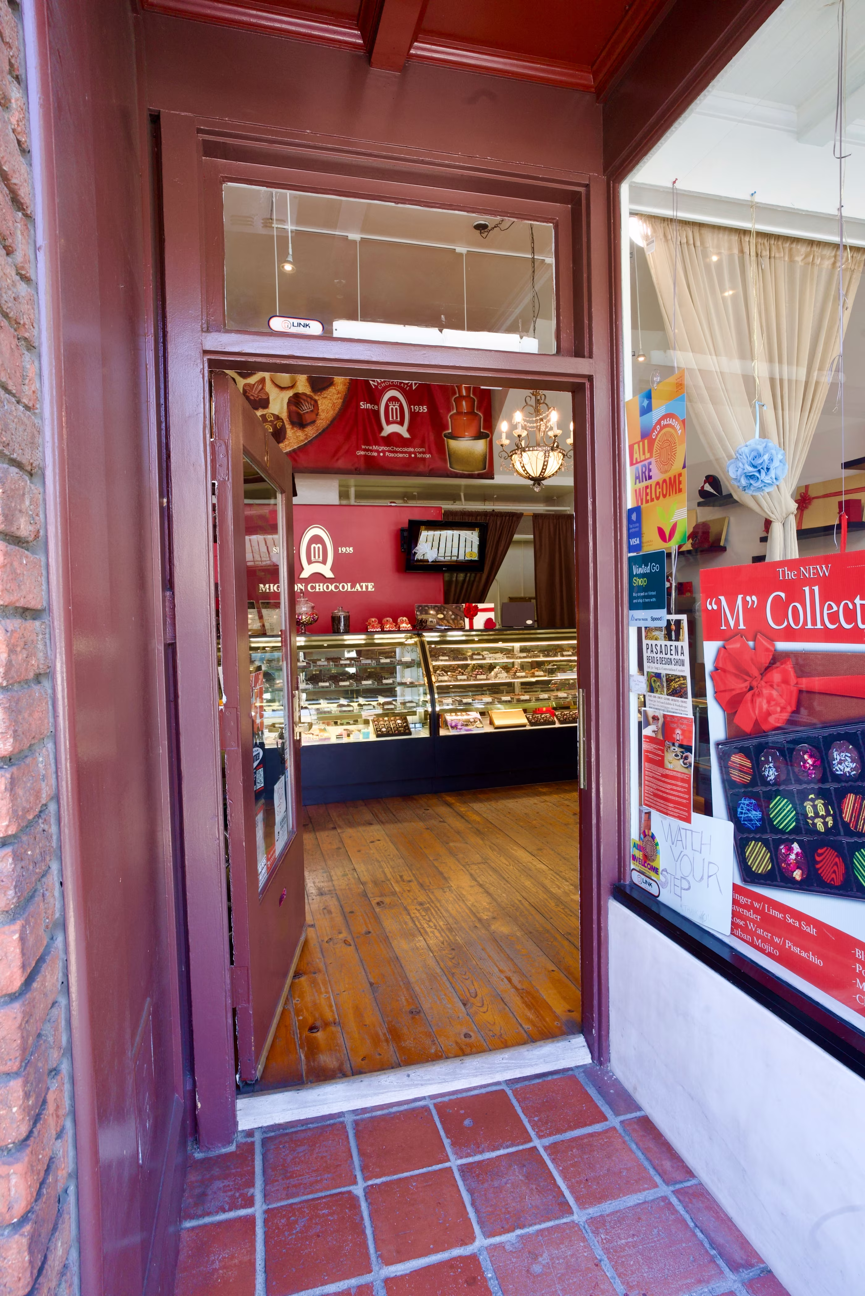 Vertical threshold view through an open burgundy storefront door toward wood flooring, illuminated display cases, chandelier, and red logo wall.