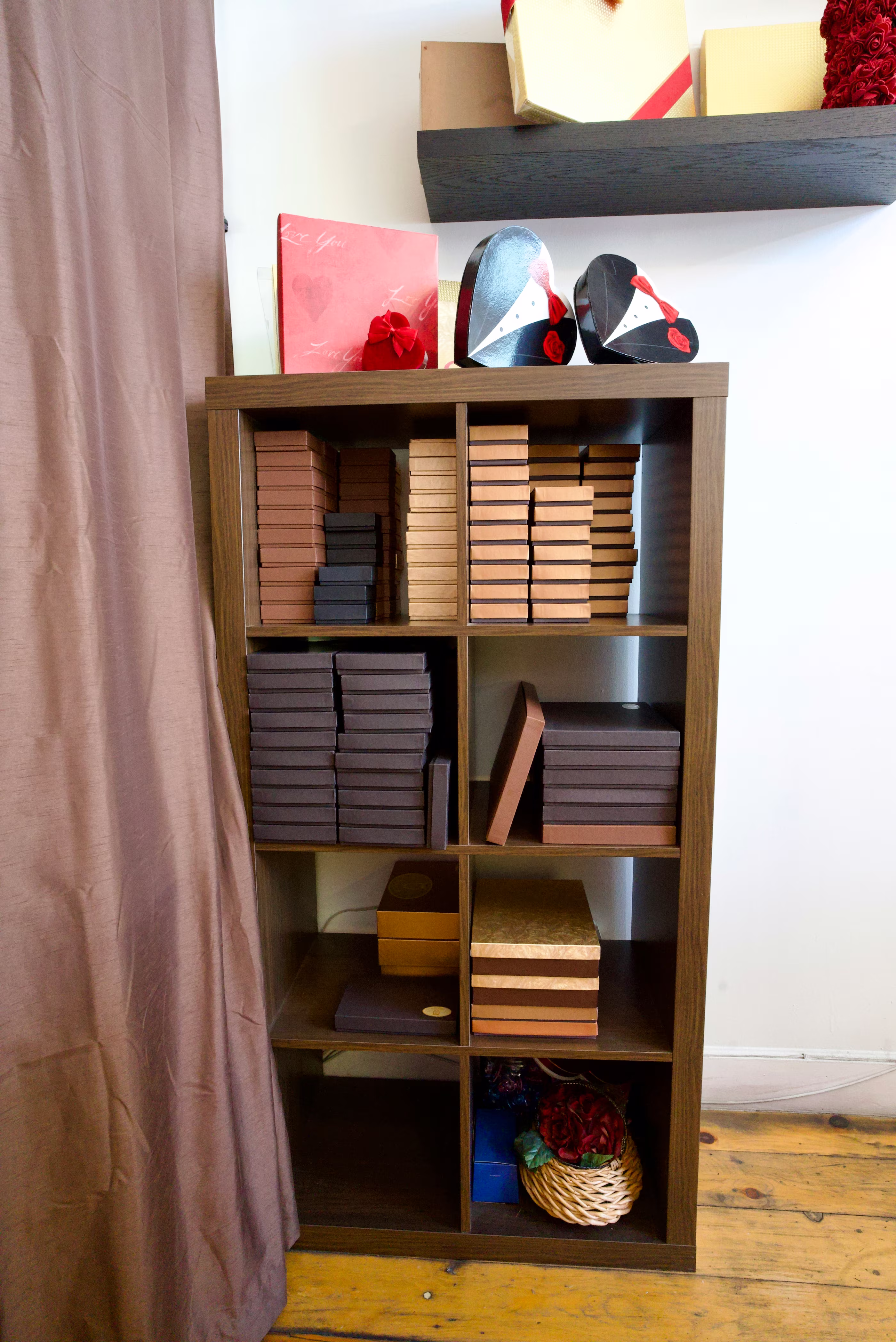 Vertical view of a dark wood cubby shelf stacked with flat gift boxes, topped by tuxedo-shaped and heart-shaped packages beside a curtain.