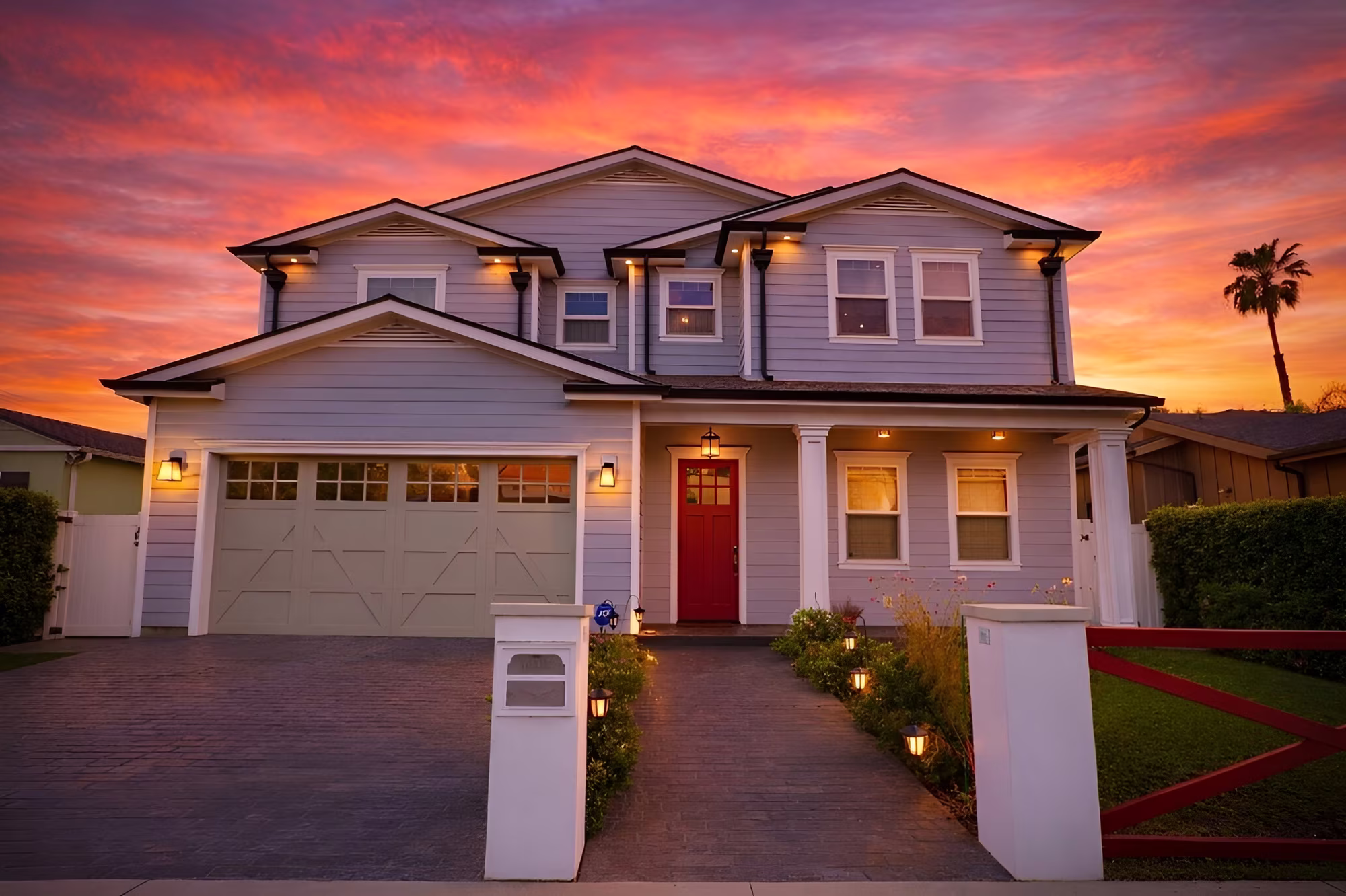 Two-story gray house with a red front door and attached garage beneath a vivid sunset sky.