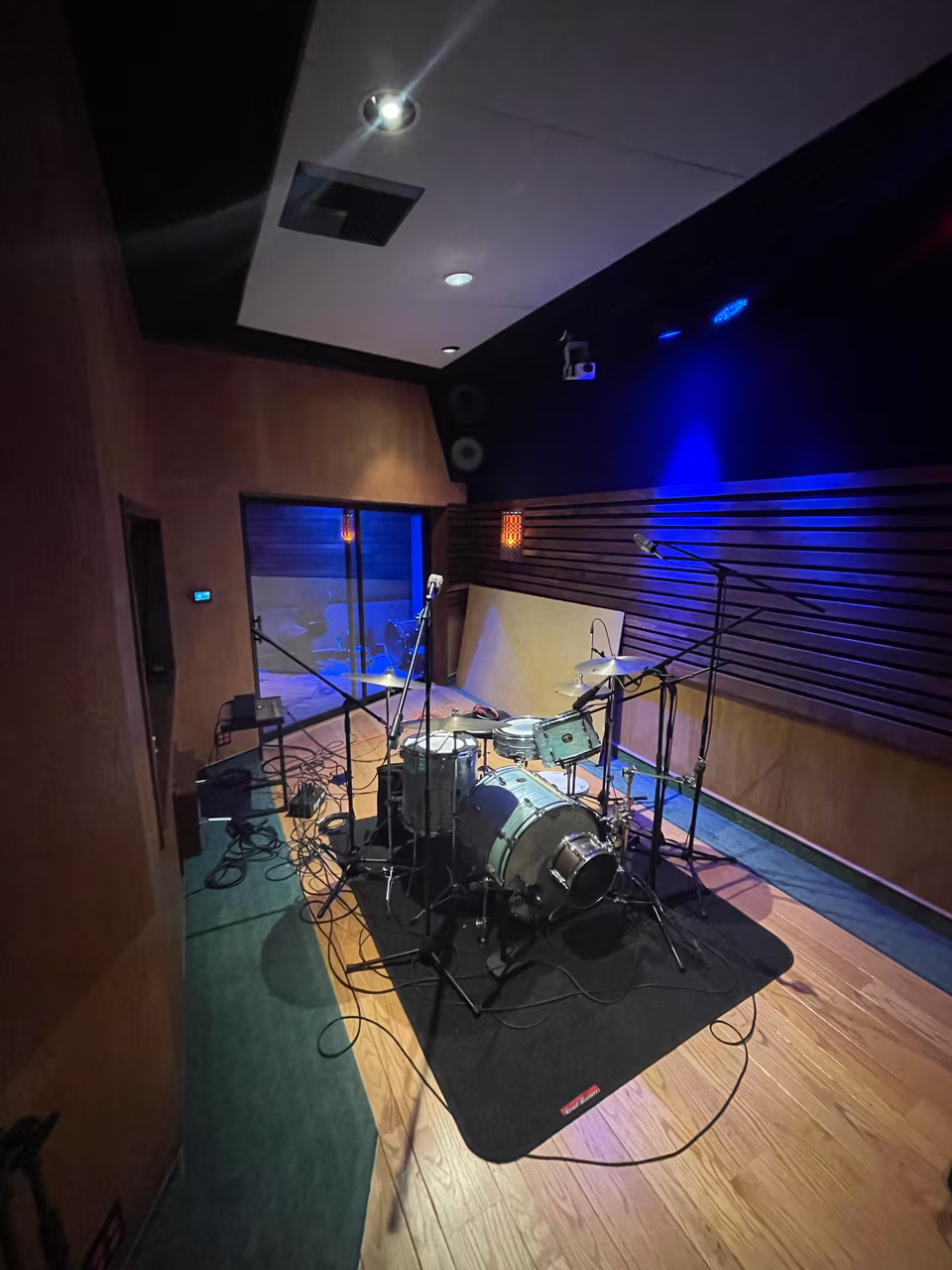 Live recording room set with a drum kit, microphones, wood flooring, and slatted acoustic walls.