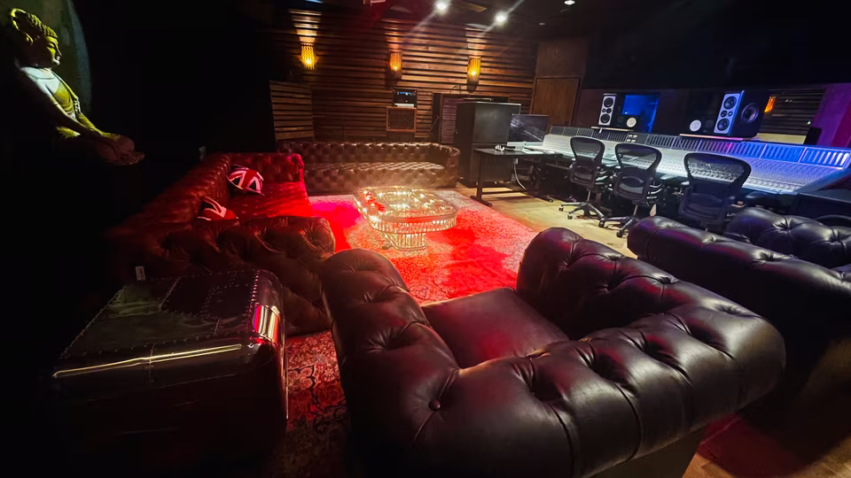 Reverse view across leather seating and a lit glass coffee table toward the mixing console and monitor speakers.