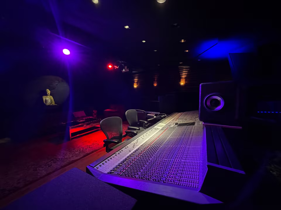 Large mixing console beneath purple and red studio lights with acoustic wood treatment and a seated statue.