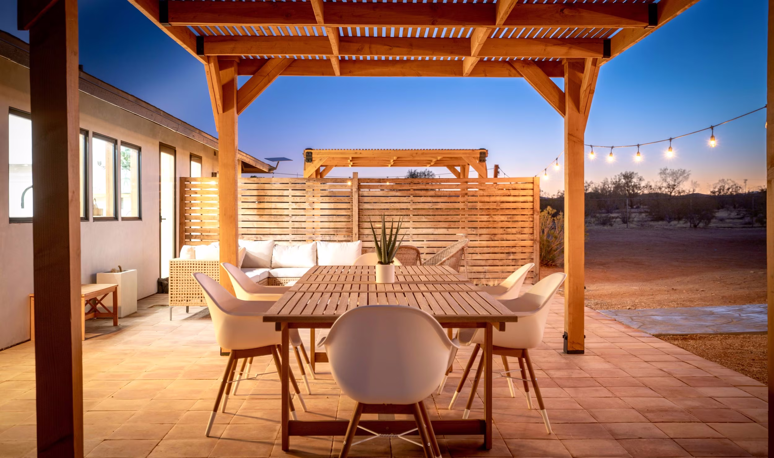 Outdoor dining table beneath a wood pergola with lounge seating and string lights at dusk.