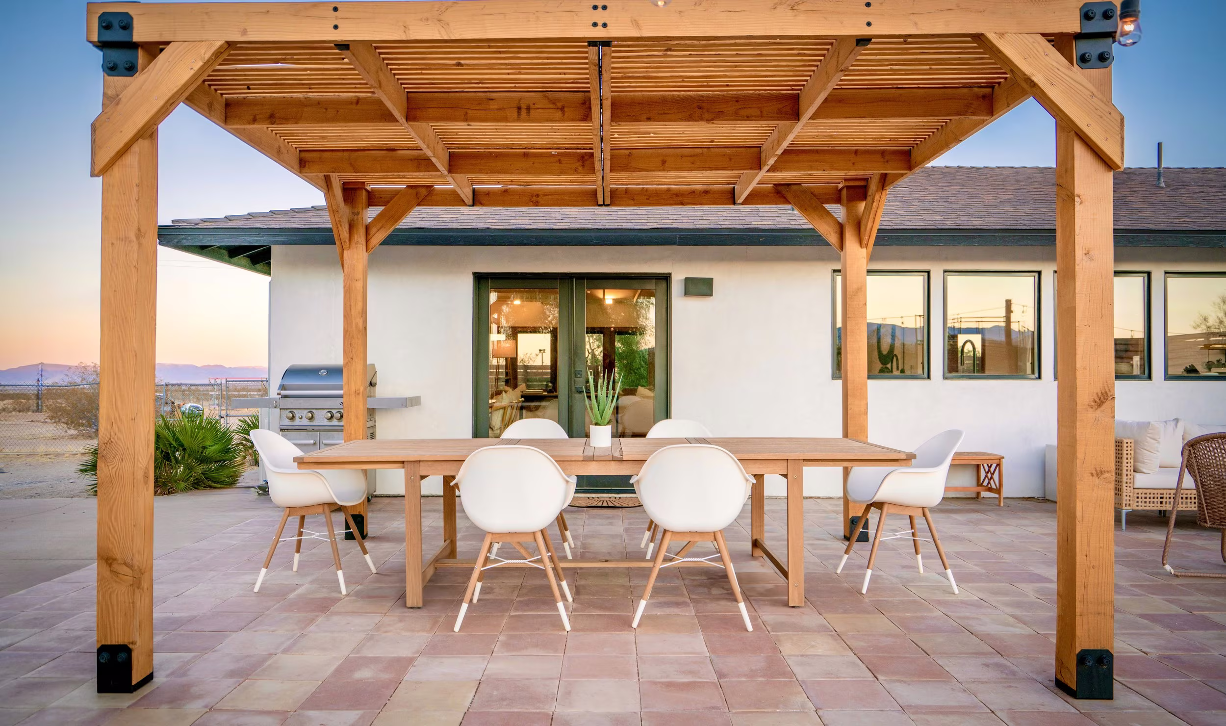 Dining table centered beneath a wood pergola with the low white house behind it.