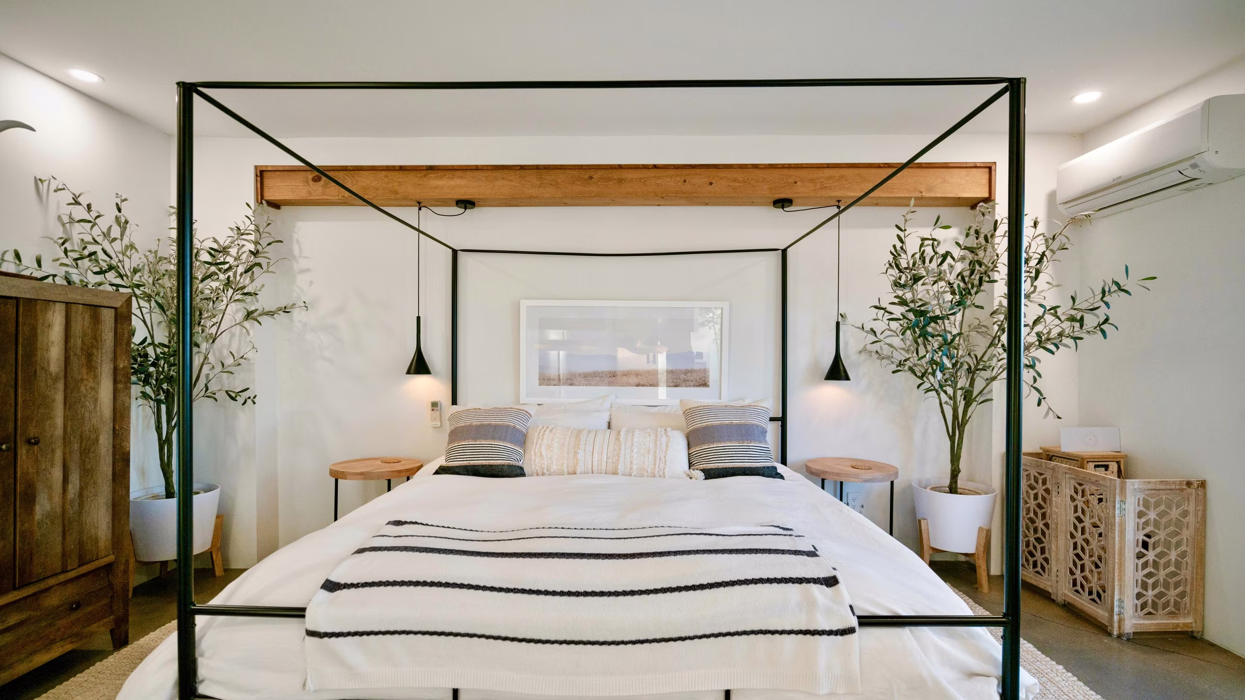 Black canopy bed with white linens between pendant lights, plants, and a rustic wood cabinet.