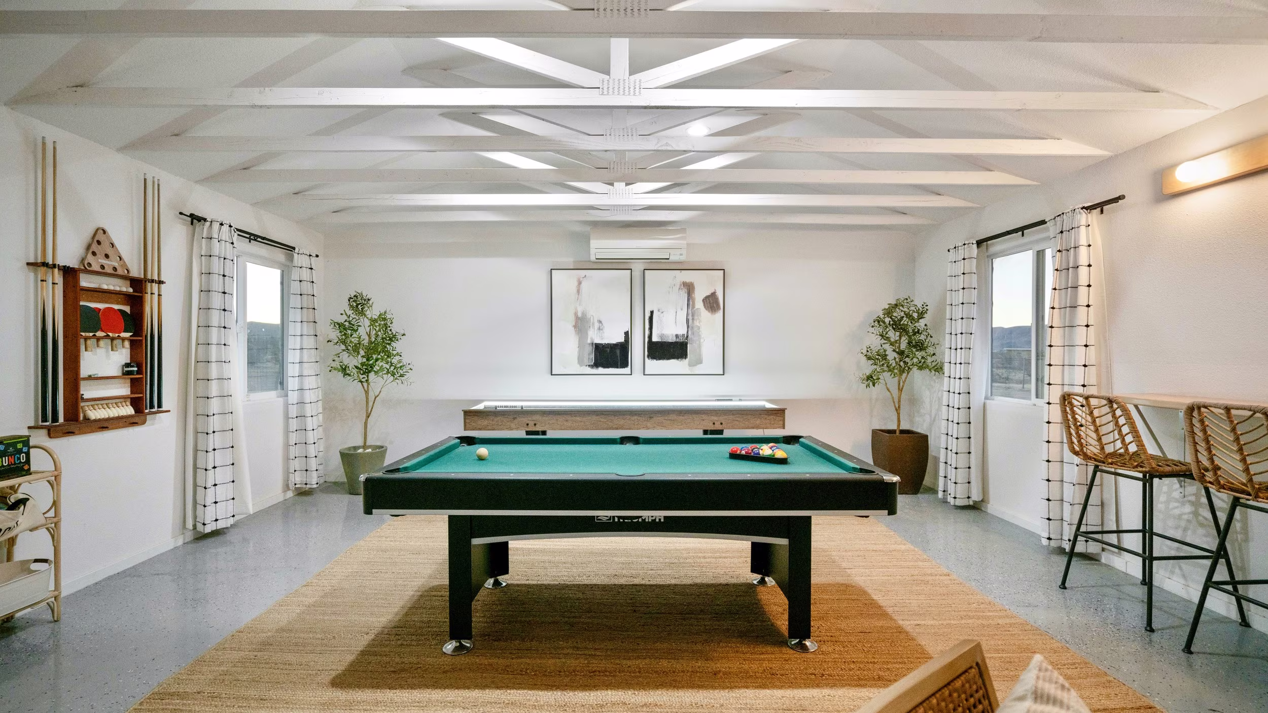 Game room with a green pool table, long shuffleboard table, white beams, and windows on both sides.