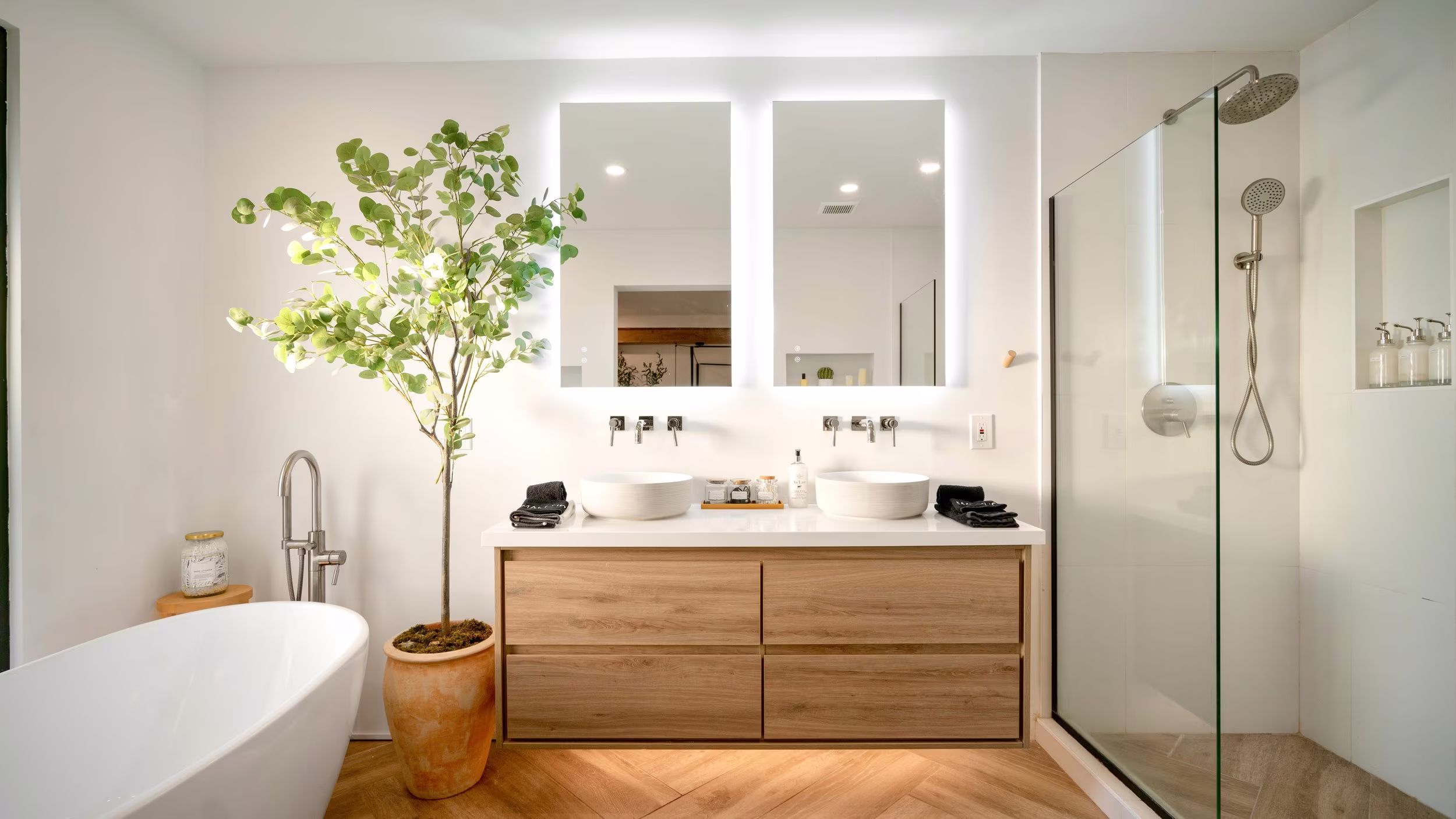 Bathroom with a double vessel-sink vanity, freestanding tub, glass shower, and herringbone wood floor.
