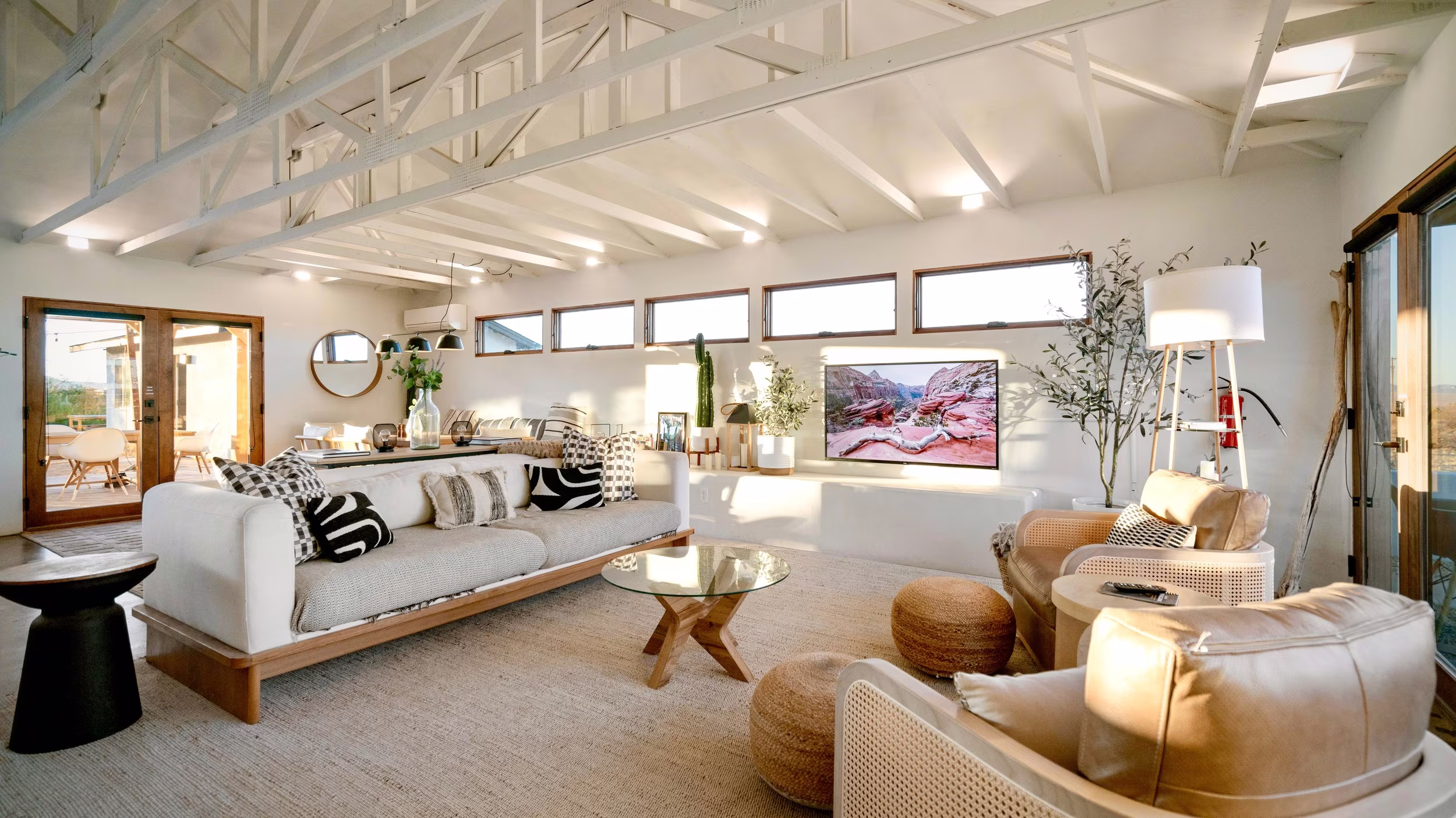 Wide living room with exposed white beams, neutral seating, television, and an open kitchen beyond.