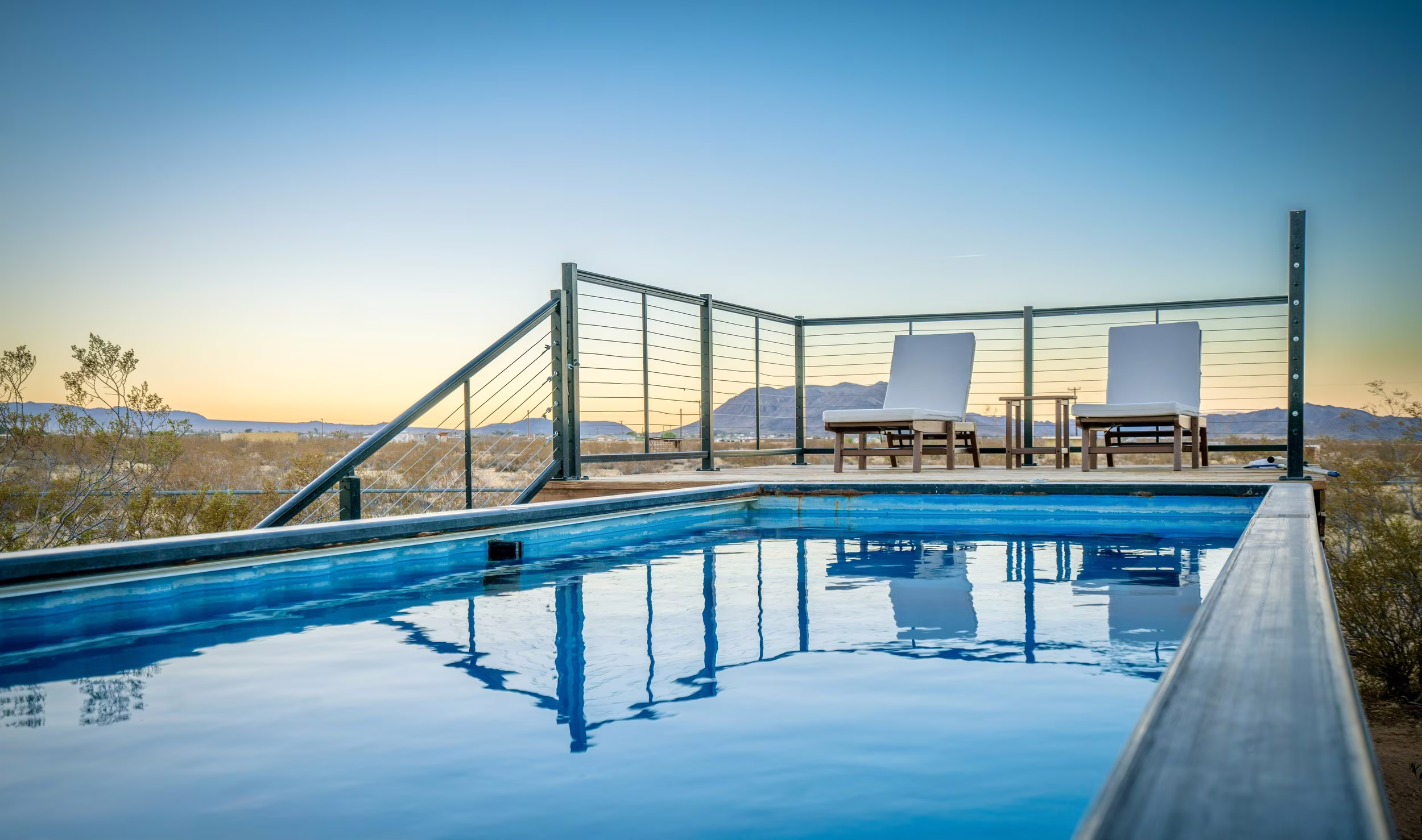Container pool reflecting the blue sky with two white loungers and distant mountains.