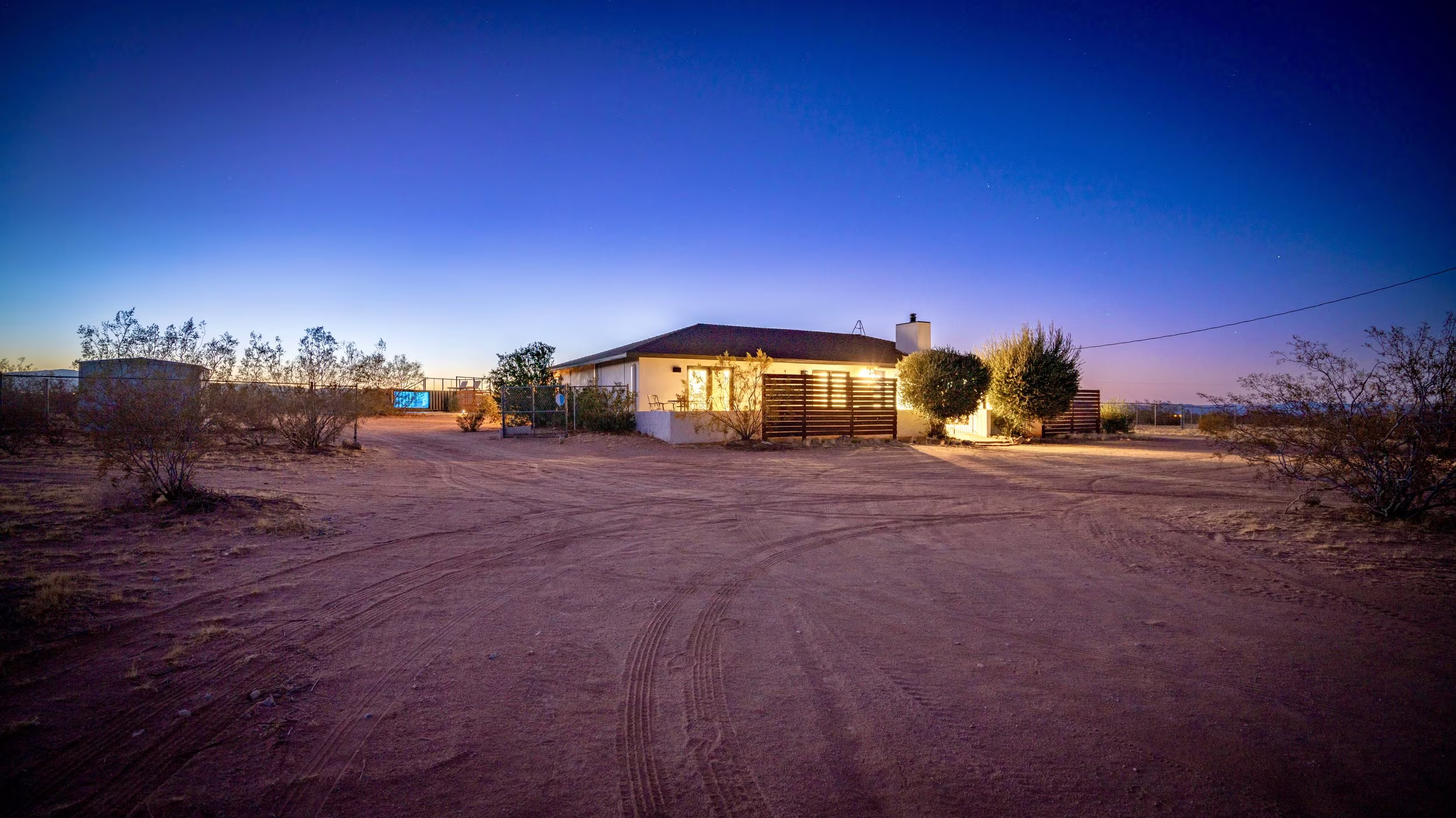 Low house glowing at dusk beyond a broad dirt drive, with the blue-lit pool visible to one side.