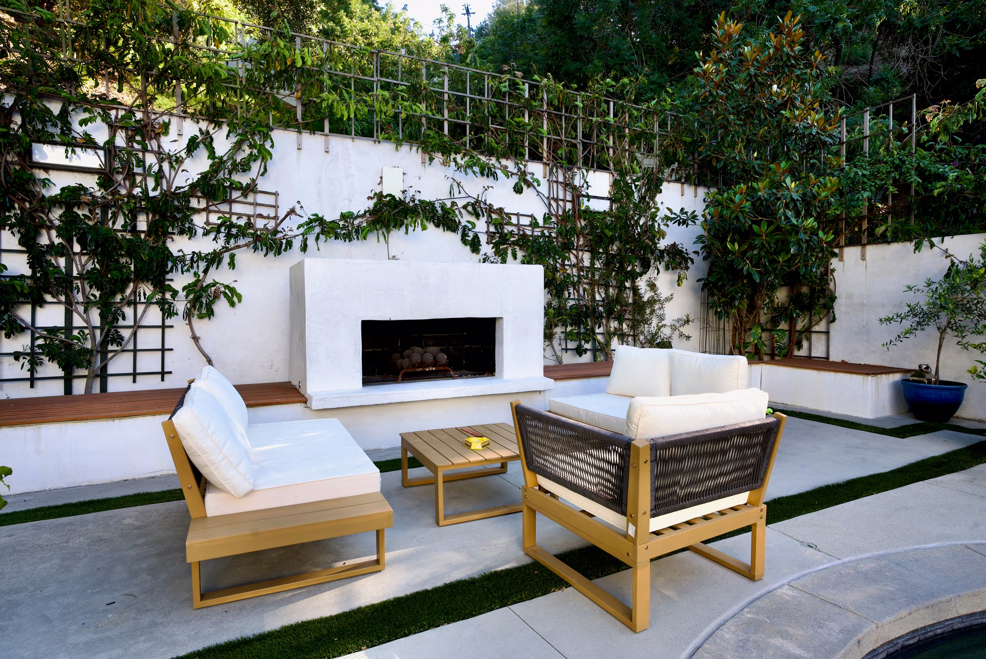 Outdoor seating arranged around a low table before a white fireplace and vine-covered trellis.