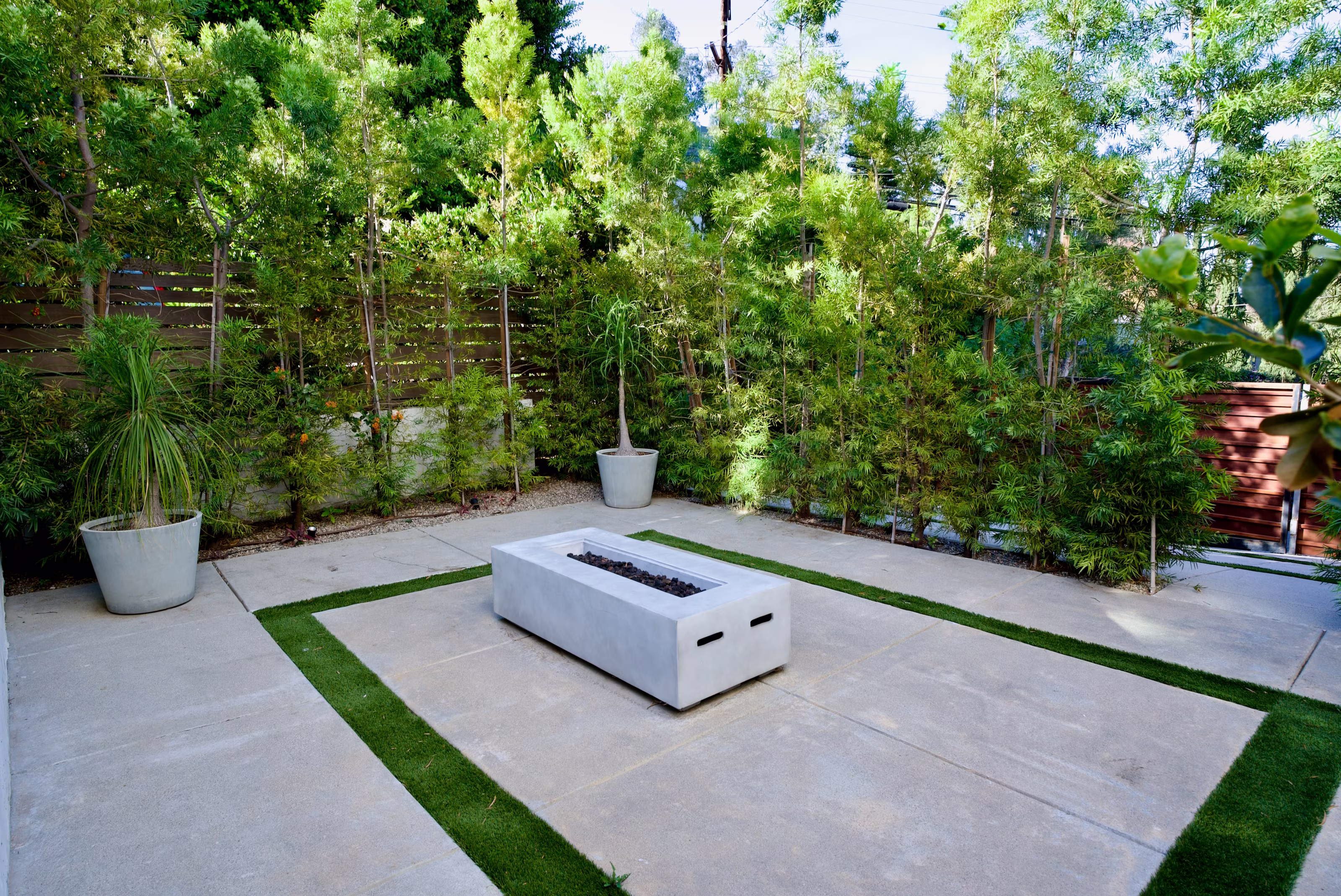 Rectangular fire pit centered on pale paving with narrow turf bands and potted palms.