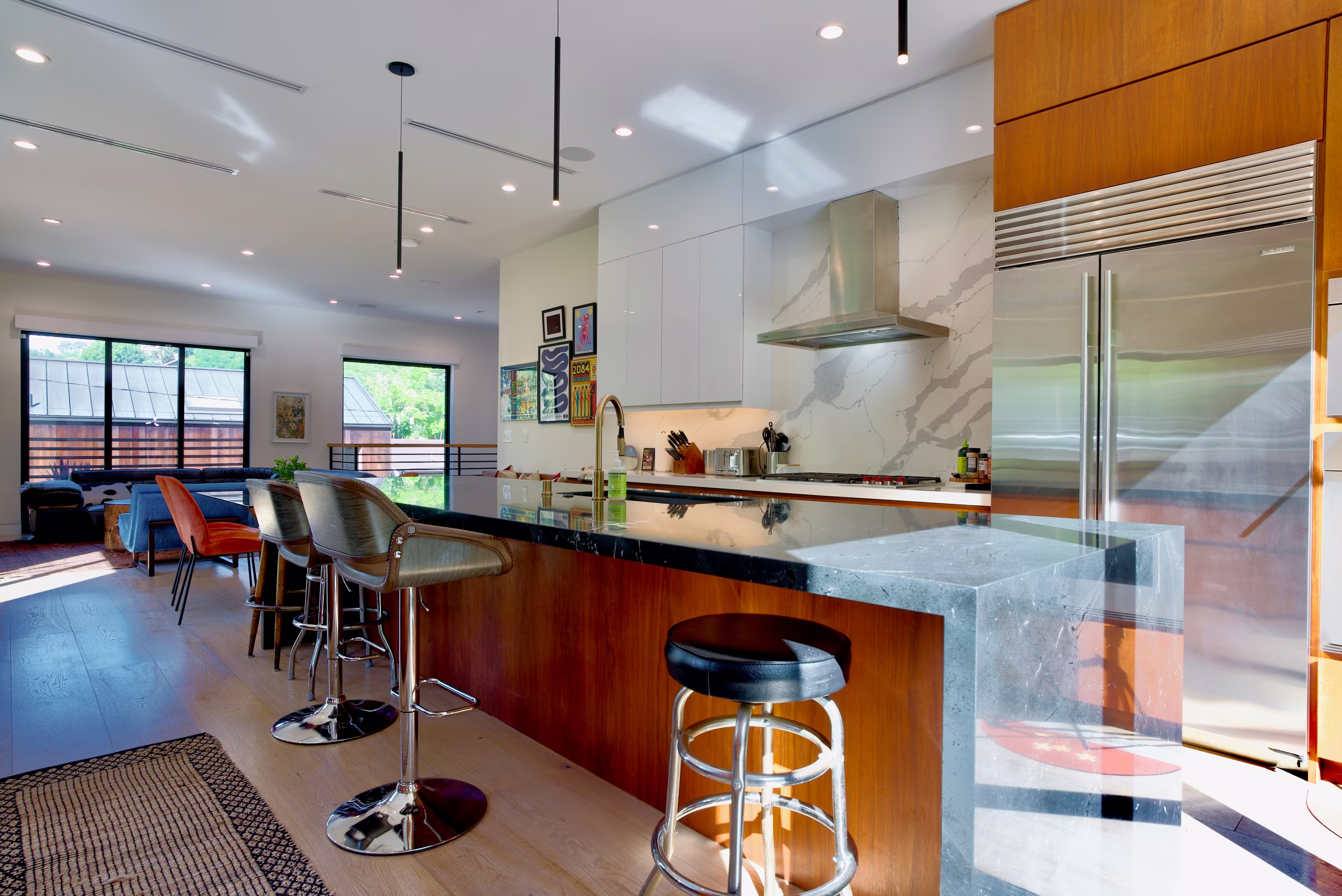 Wide kitchen view showing a waterfall-edge stone island, stainless refrigerator, and adjacent lounge.