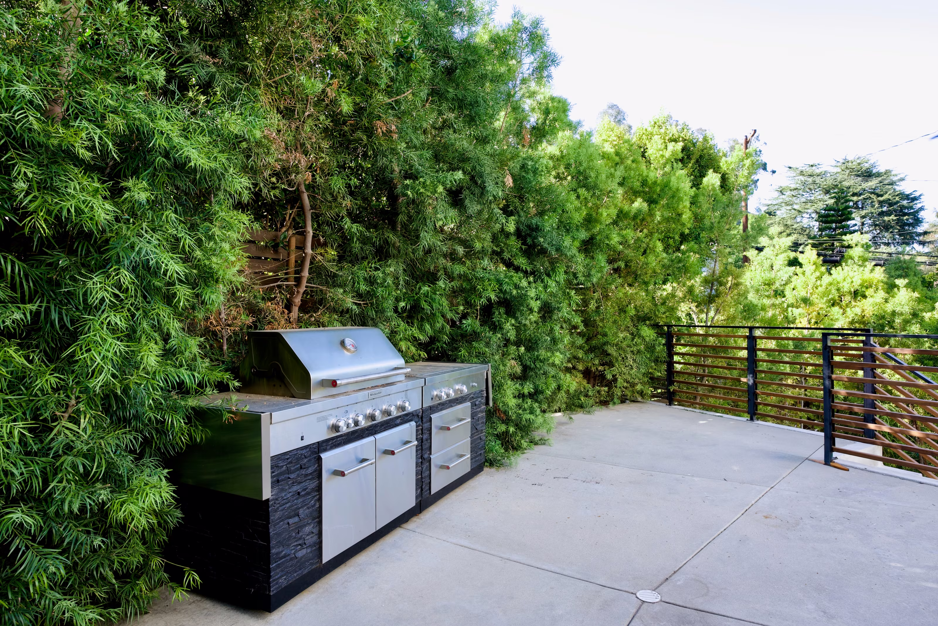 Stainless outdoor grill and prep cabinets beside a slatted gate and tall hedges.
