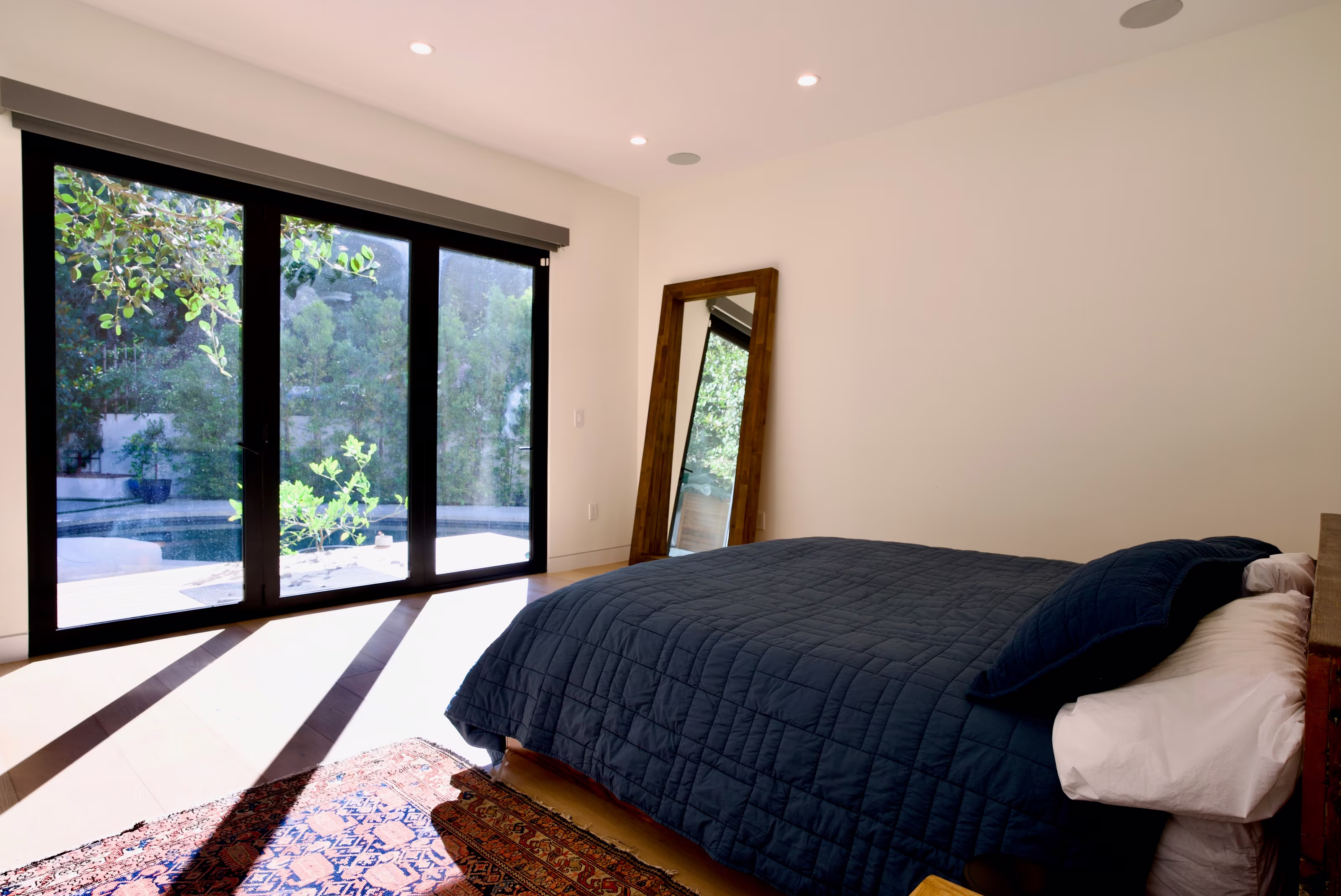Primary bedroom with dark blue bedding, garden-facing sliding glass doors, and a leaning wood-framed mirror.