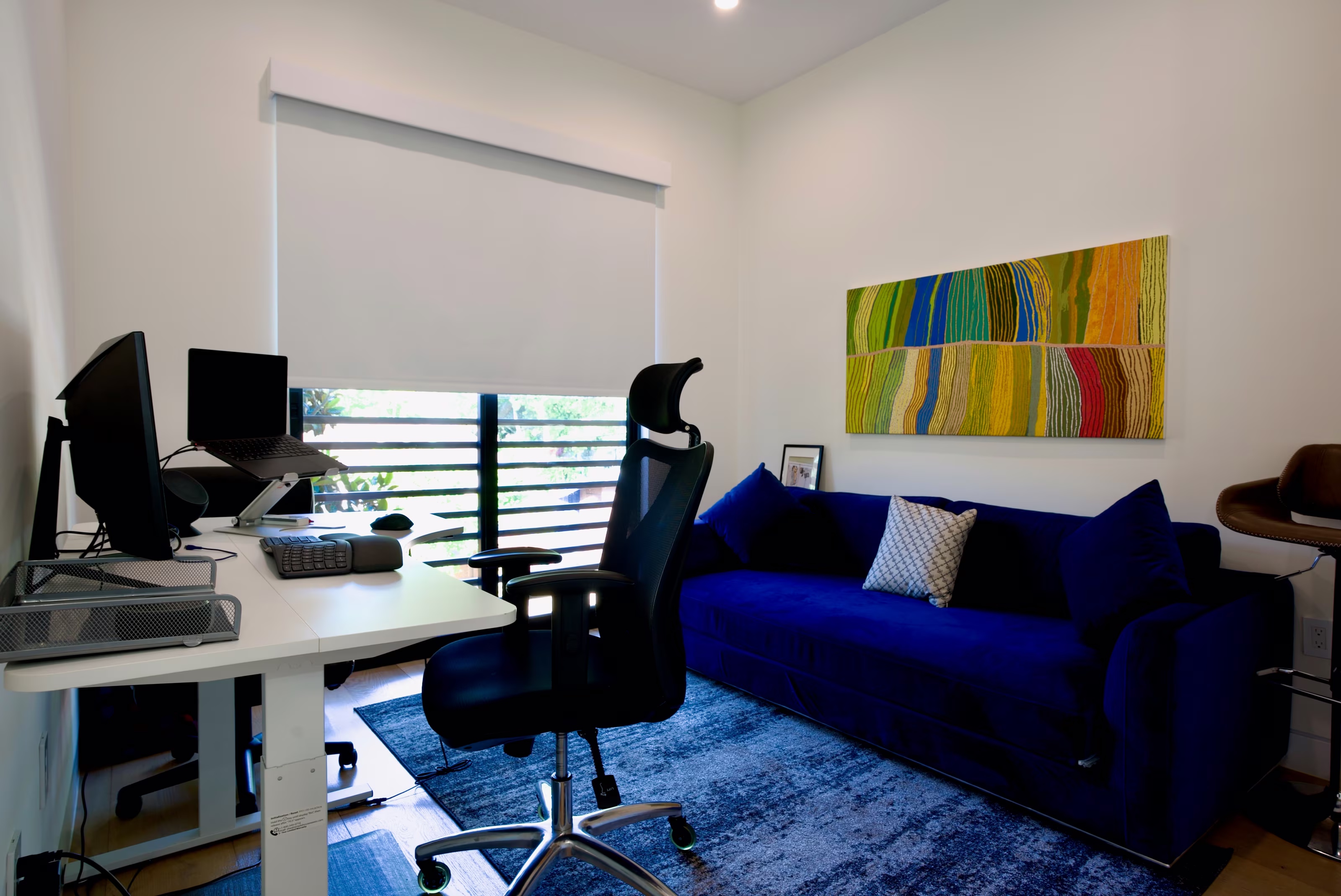 Office with dual-monitor desk, black ergonomic chair, blue sofa, striped artwork, and balcony window.