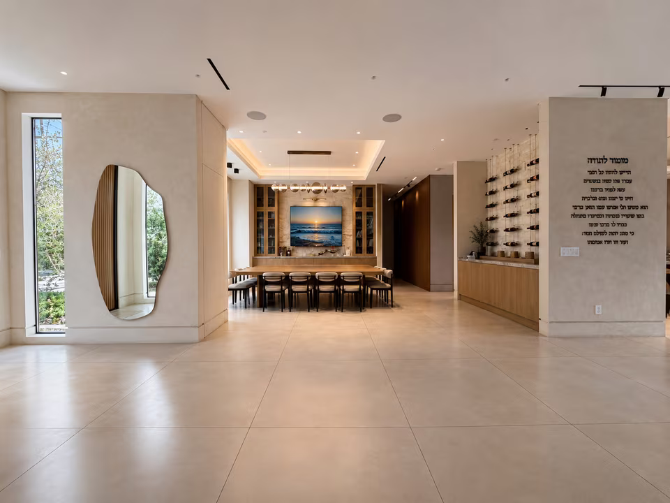 Open entry and dining area with a long table, sculptural lighting, warm wood floors, and a skylit stair beyond.