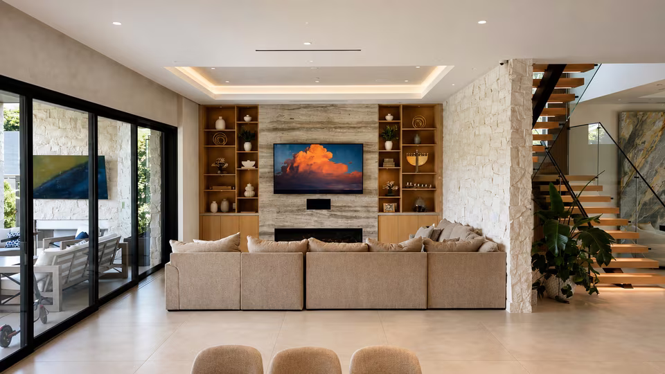 Bright living room with pale seating, warm wood floors, a stone fireplace wall, and wide glass doors opening toward the pool.