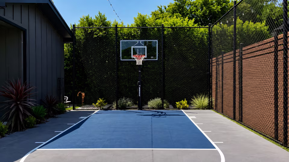 Private outdoor sport court with basketball hoops, painted game lines, tall fencing, and trees beyond.