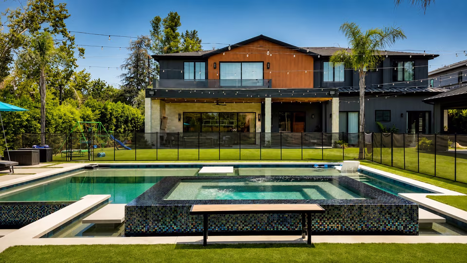 Wide rear view across a large swimming pool and raised spa toward the two-story home and covered outdoor lounge.