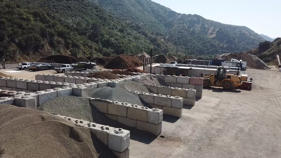 Wide yard view with a front loader, parked vehicles, material bays, and equipment beneath open sky.