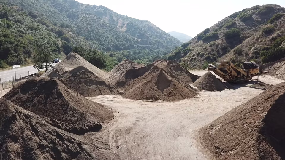 Broad aggregate field with pale rock piles, heavy machinery, screening equipment, and rugged ground texture.