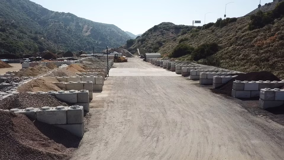 Elevated view down a broad dirt lane between aggregate piles, heavy equipment, and outdoor storage bays.