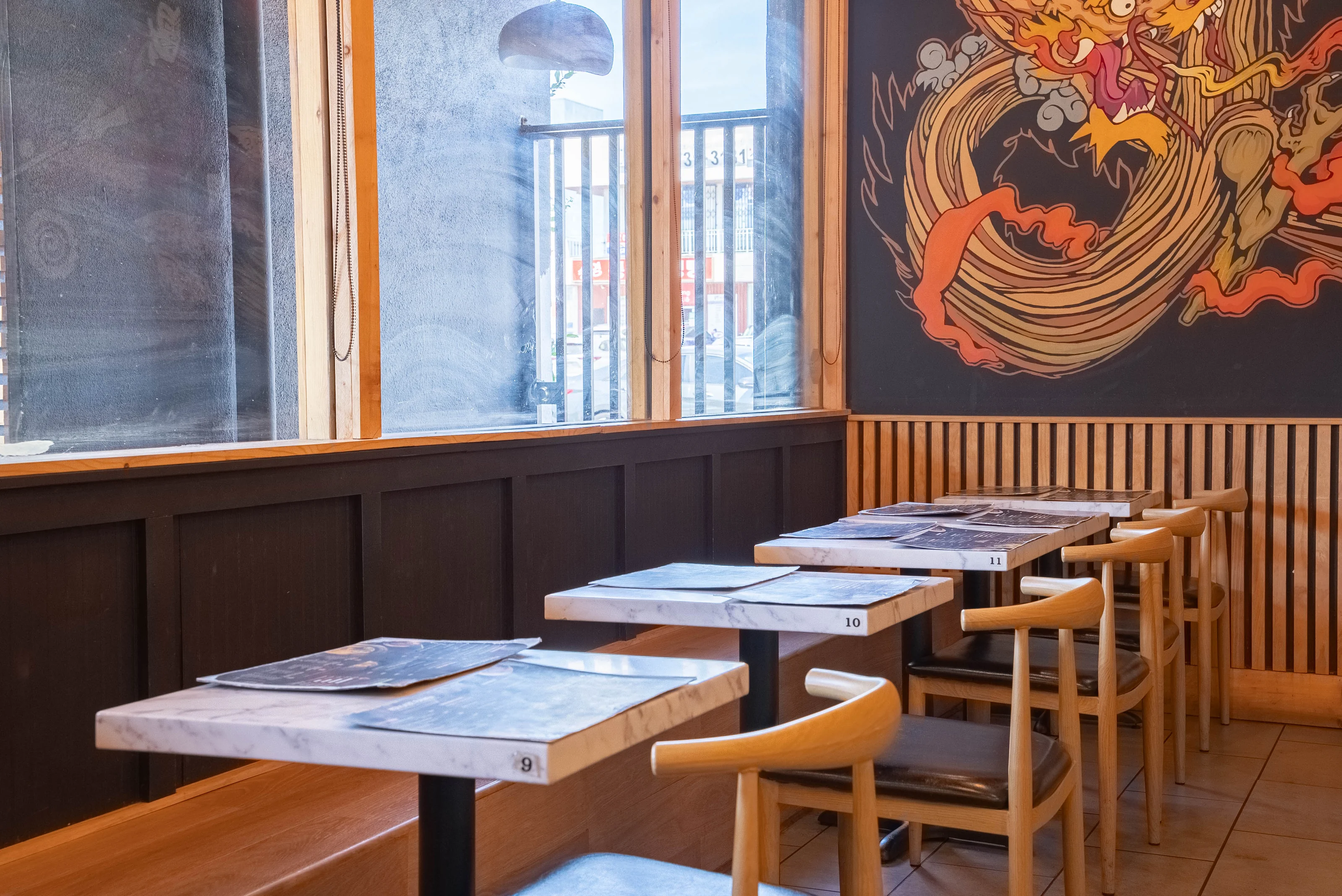 Window-side banquette tables with pale wood chairs beneath a partial dragon mural and warm slatted wall detailing.